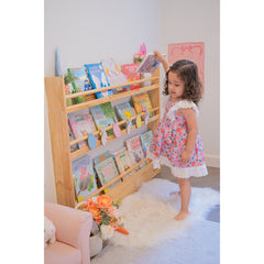 A young girl placing books on a JORY 3 — Front-Facing Bookshelf in a bright, cozy reading corner.