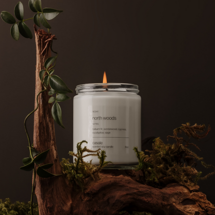 NORTH WOODS SINGLE WICKED SOY CANDLE (8oz / 226g) lit on natural wood with greenery, helping families thrive with tools to calm.