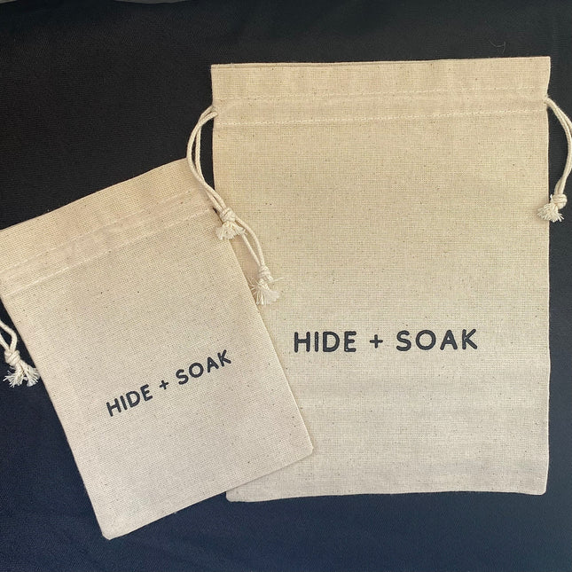 Two natural cotton drawstring bags labeled Hide + Soak Cotton Bath Bags – Small & Large | Reusable on black background