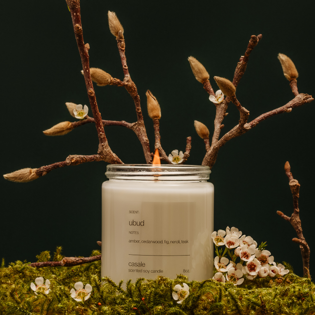 UBUD SINGLE WICKED SOY CANDLE (8oz / 226g) lit among delicate white flowers and moss, helping families thrive.