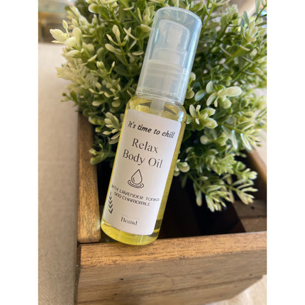 Clear bottle of Relax Body Oil Massage and Moisturise with pump, displayed by green leafy plant in wooden box
