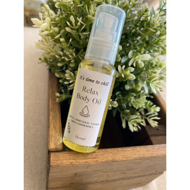 Clear bottle of Relax Body Oil Massage and Moisturise with pump, displayed by green leafy plant in wooden box