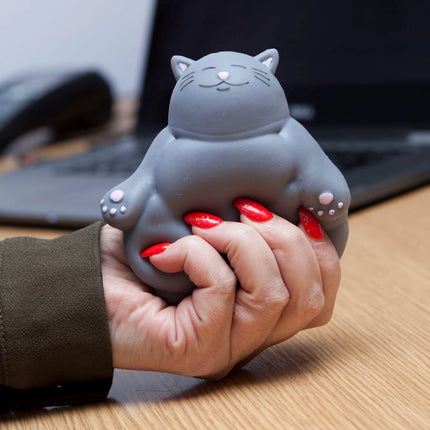 Hand squeezing a gray cat-shaped Stress Toy - Kalma Kitty on a desk with laptop in the background helping families thrive.