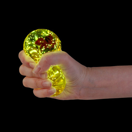 Hand squeezing glowing yellow Smooshos Super Squeeze Diamond Sphere GID Dino sensory squishy toy with dinosaur inside.