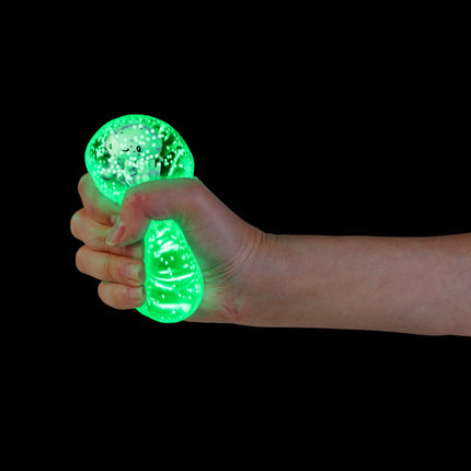 Hand squeezing glowing green Smooshos Super Squeeze Diamond Sphere GID Sea Animal sensory fidget toy against black background