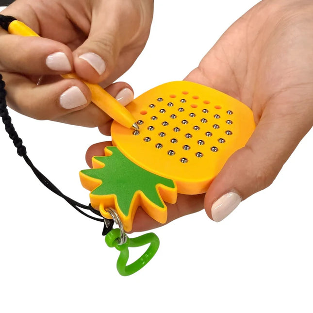 Hands holding a bright yellow and green Pineapple Magnetic Pad Keyring, using a magnetic stylus to move metal balls on the pad surface.