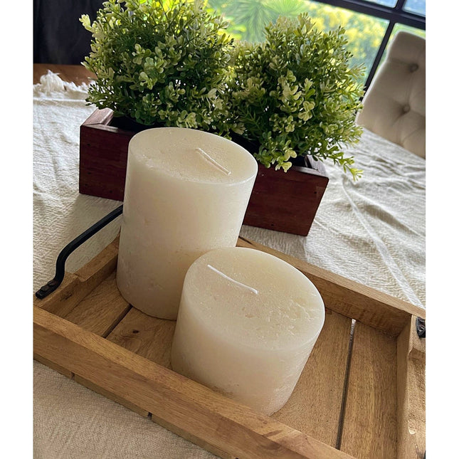 Two off white pillar candles rustic relic design on wooden tray with green plant centerpiece in background