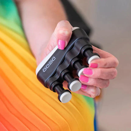 Hand holding a black and white Hand & Finger Grip Exerciser & Fidgeting Sensory Tool with pink nail polish against a colorful background.