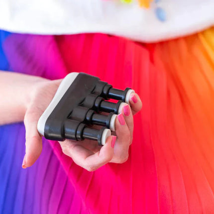 Hand holding a black and white Hand & Finger Grip Exerciser & Fidgeting Sensory Tool against a colorful pink, red, and orange background.