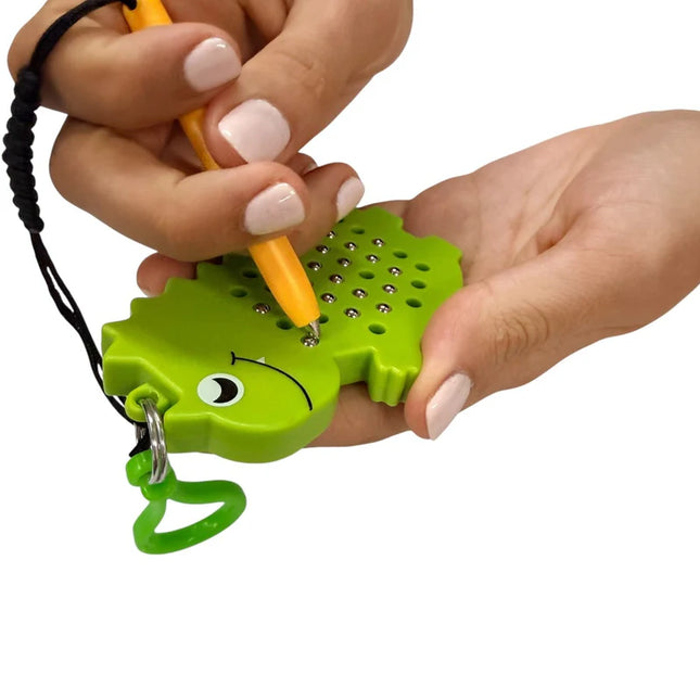 Hand holding a green dinosaur-shaped magnetic pad keyring with a stylus pen lifting small metal balls from the surface, demonstrating the Dinosaur Magnetic Pad Keyring.