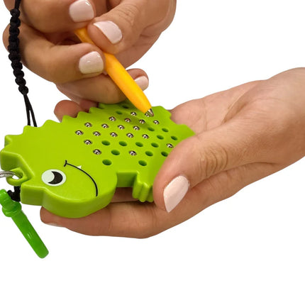 Hands holding a green dinosaur-shaped toy with metal balls being moved by a yellow magnetic pen, featuring the Dinosaur Magnetic Pad Keyring.