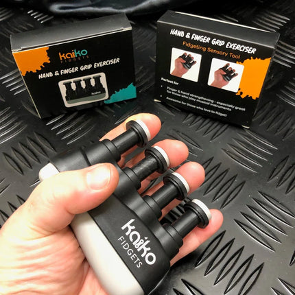 Hand holding a black and white Hand & Finger Grip Exerciser & Fidgeting Sensory Tool with product packaging visible in the background on a textured black surface.