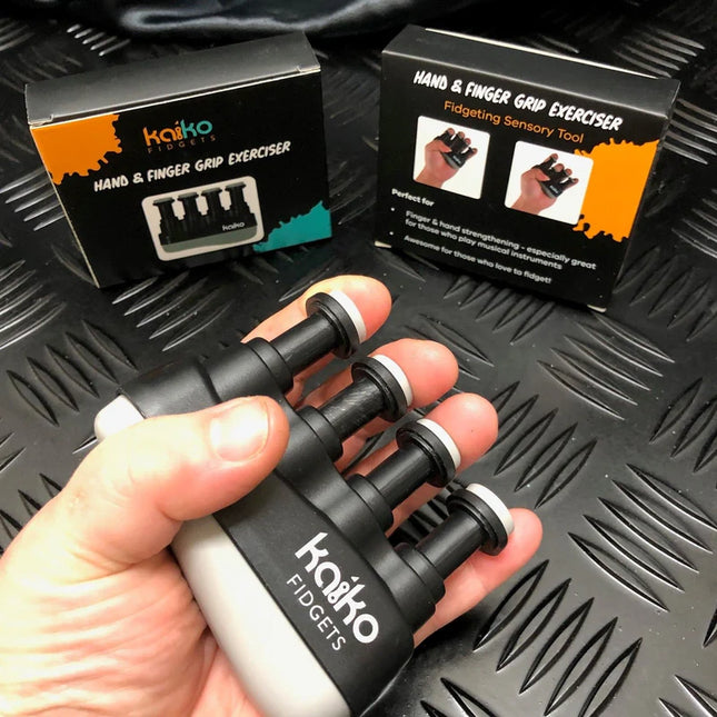 Hand holding a black and white Hand & Finger Grip Exerciser & Fidgeting Sensory Tool with product packaging visible in the background on a textured black surface.