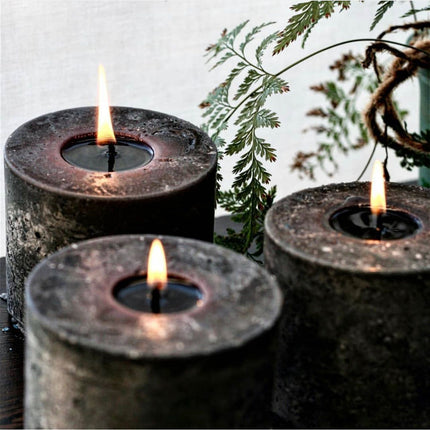 Charcoal Black Pillar Candle Stone Relic Finish trio with flickering flames beside delicate green foliage, calming ambiance.