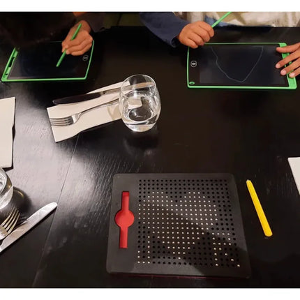 Two children using green styluses on Magnetic Pad - Original Rectangle Design tablets with a perforated black board and yellow stylus on a dark table alongside glassware and cutlery.