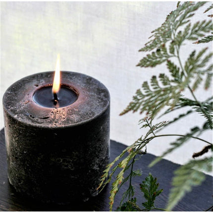 Charcoal Black Pillar Candle Stone Relic Finish burning softly beside green fern branches, calming home decor.