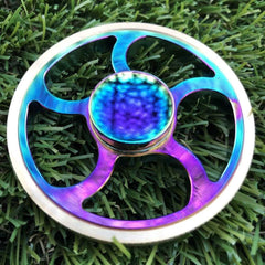 Flywheel Spinner Fidget in iridescent blue and purple metal resting on green grass background.