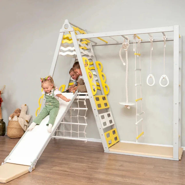 Children playing on a white and yellow Wooden Teepee Playground with slide, climbing ropes, and gymnastic rings indoors.